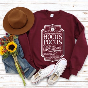 Peut inclure: Un sweat-shirt bordeaux avec un motif blanc d'une étiquette d'apothicaire vintage qui dit "The Sanderson Sisters Hocus Pocus Apothecary" avec le texte "Fine Potions, Mystical Brews, Magic Elixers, Tonics & Tinctures" en dessous.