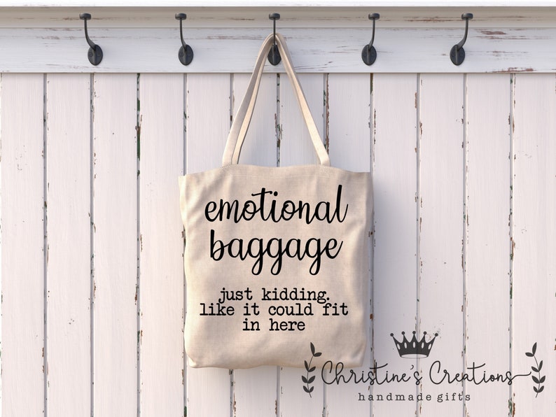 May include: A white canvas tote bag with black text that reads "emotional baggage just kidding, like it could fit in here". The bag is hanging on a white wooden hook rack. The text "Christine's Creations handmade gifts" is also visible on the bag.