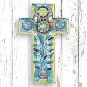 A handcrafted wall-hanging cross made from natural oak veneer, featuring an intricate collage design in sunshine yellow and sky blue. The central "I am the vine" motif features a leafy tree with white blossoms, with theological text snippets like "Holy Spirit," "God," "Heaven," and "I am Jesus" collaged throughout. The design is bright and uplifting, shown against a white rustic wood-plank background.