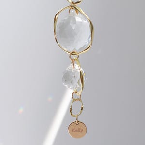 May include: A gold-colored suncatcher with three clear, faceted crystal orbs suspended by gold rings and chains. A small, round gold charm at the bottom is engraved with the name "Kelly".