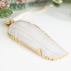 May include: A white feather pendant with a gold trim. The feather is carved with a detailed pattern and hangs from a gold chain.