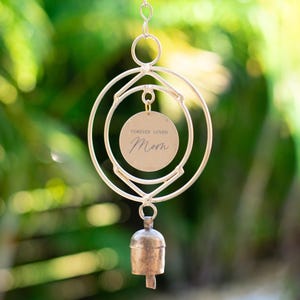 May include: A silver wind chime with a circular design and a bell at the bottom. A small round tag hangs from the center of the chime with the text "Forever Loved Mom".
