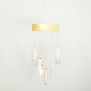 May include: A decorative wind chime featuring a rectangular gold-colored metal plate suspended by a gold chain. Six white and pink feather-shaped pendants with gold trim hang below. The wind chime is designed to be hung indoors or outdoors.