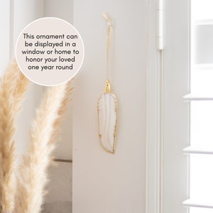 May include: A white feather ornament with gold accents hangs from a hook on a white wall. The ornament can be displayed in a window or home to honor a loved one year round.