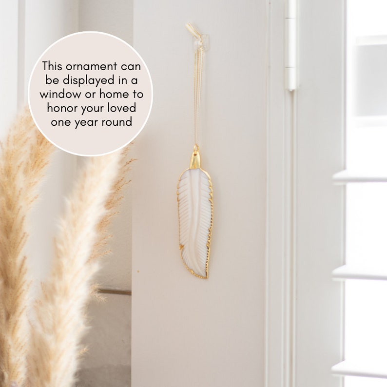 May include: A white feather ornament with gold accents hangs from a hook on a white wall. The ornament can be displayed in a window or home to honor a loved one year round.