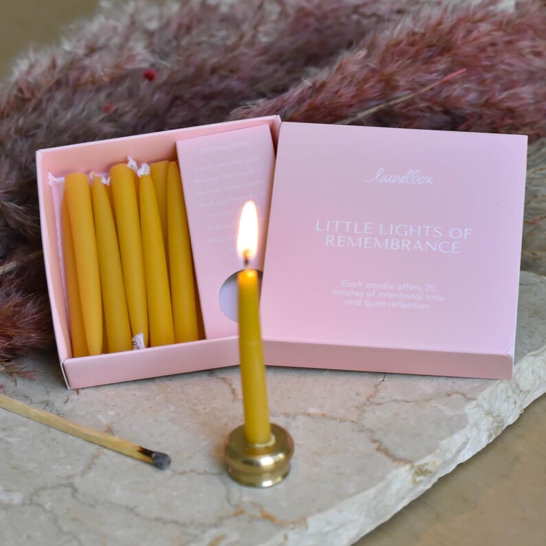 May include: A lit yellow candle in a gold holder next to a box of unlit yellow candles. The pink box reads "Little Lights of Remembrance". A burnt matchstick lies nearby. The background features dried, feathery, pinkish-brown plants.