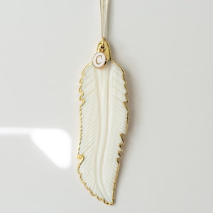May include: A white feather ornament with a gold trim and a gold charm with the letter "C" on it.