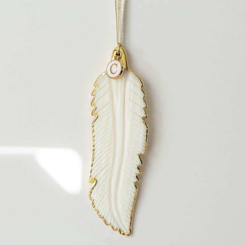 May include: A white feather ornament with a gold trim and a gold charm with the letter "C" on it.