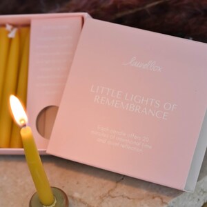 May include: A lit yellow candle in a brass holder with a pink box labeled "LITTLE LIGHTS OF REMEMBRANCE." The box contains several yellow candles. Text on the box states each candle offers 20 minutes of intentional time.