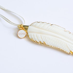 May include: A white feather pendant necklace with a gold trim and a small round charm that says "MOM" in pink lettering.