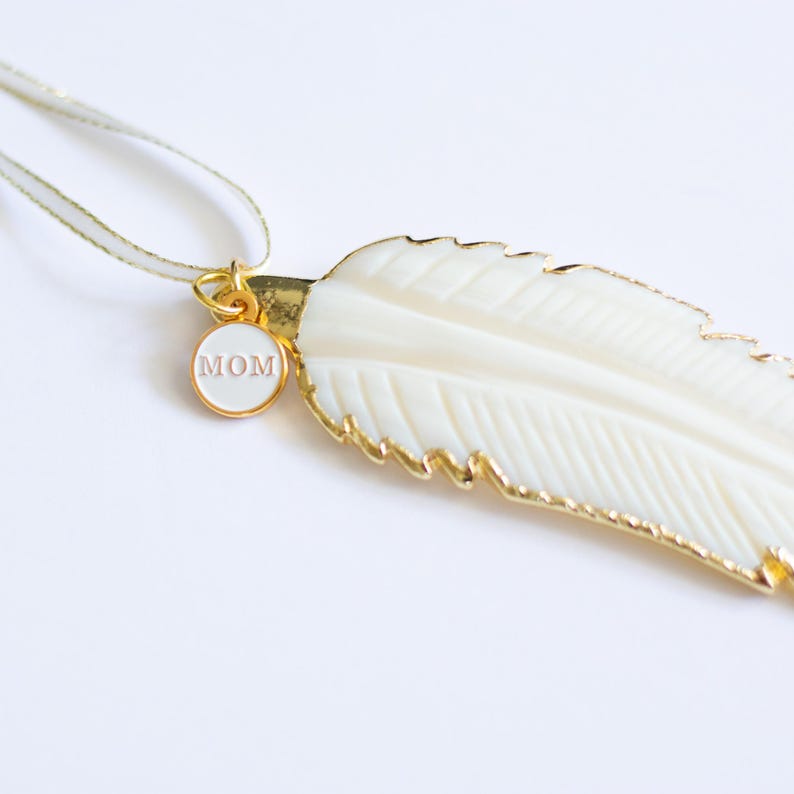 May include: A white feather pendant necklace with a gold trim and a small round charm that says "MOM" in pink lettering.