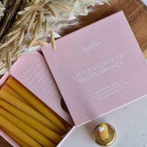 May include: A pink box with the text "LITTLE LIGHTS OF REMEMBRANCE" contains several yellow candles. The box also includes instructions and a small candle holder. Dried flowers are in the background.