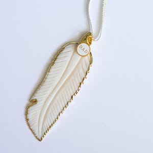 May include: A white feather pendant with a gold trim and a small round tag that says "DAD". The pendant is hanging from a white ribbon.