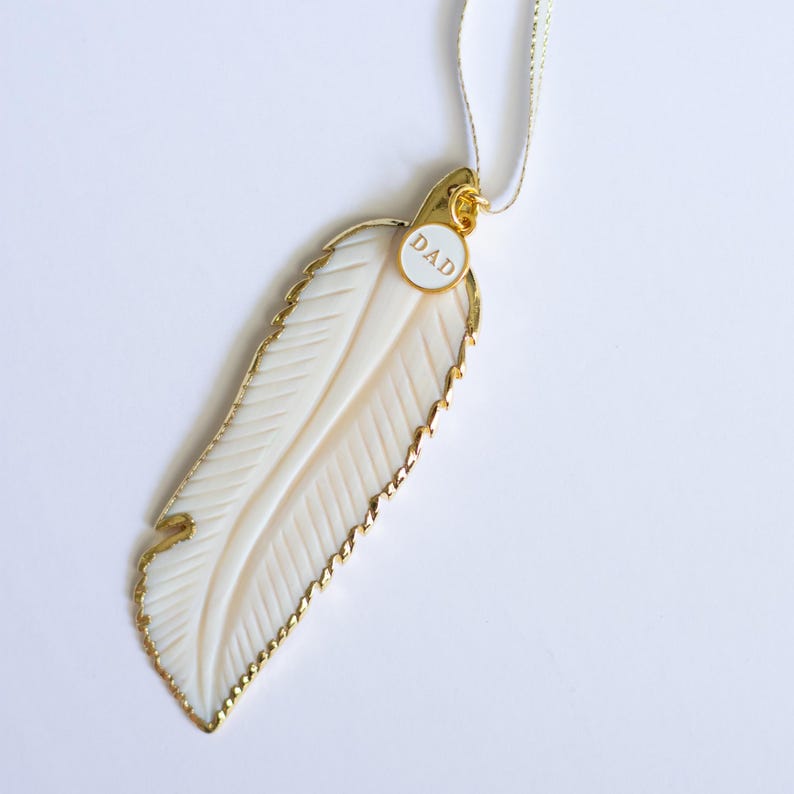 May include: A white feather pendant with a gold trim and a small round tag that says "DAD". The pendant is hanging from a white ribbon.