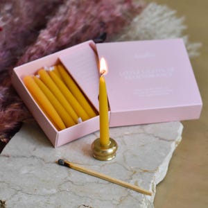 May include: A lit yellow candle in a brass holder, with a box of unlit candles and a match. The box is pink and reads "Little Lights of Remembrance". The candles are a warm yellow color, and the match is unlit.