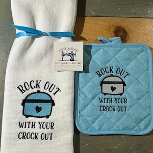 May include: A white kitchen towel and a blue pot holder with the text "Rock Out With Your Crock Out" and a stylized image of a slow cooker. The text "Custom Embroidery Red Rock Crafts Wy" is on a white tag.