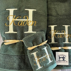 May include: A set of three green towels with a white embroidered letter H and the name Kaden in gold thread. The towels are tied with a gold ribbon. The tag reads "Custom Embroidery Red Rock Crafts Wy."