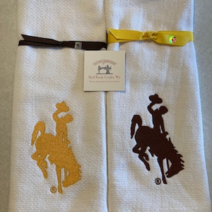 May include: Two white kitchen towels with embroidered brown and gold Wyoming bucking horse logos. The towels are tied with brown and yellow ribbon. The towels have a label that reads "Custom Embroidery Red Rock Crafts Wy Custom Embroidered Towels and More".