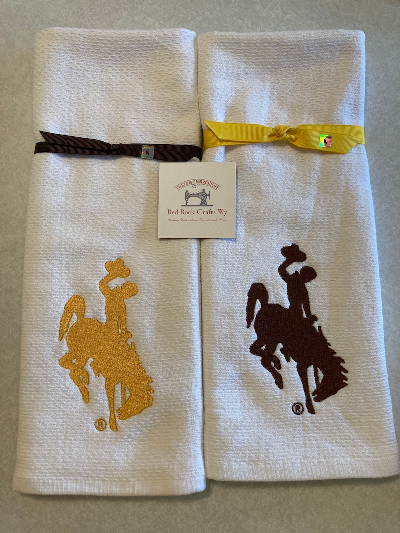 May include: Two white kitchen towels with embroidered brown and gold Wyoming bucking horse logos. The towels are tied with brown and yellow ribbon. The towels have a label that reads "Custom Embroidery Red Rock Crafts Wy Custom Embroidered Towels and More".