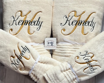 May include: Set of four cream-colored towels with gold embroidery. The towels are folded and arranged in a decorative way. The embroidery says "Kennedy" in a cursive font.