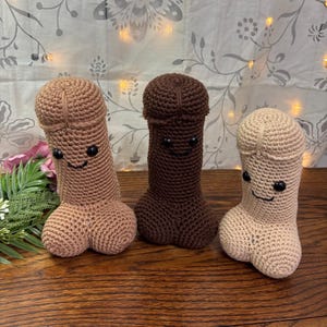 May include: Three crocheted phallus dolls in varying shades of brown and beige. Each doll has a smiling face with black button eyes and a stitched mouth. The dolls are arranged on a wooden surface.