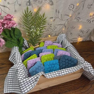 May include: A wooden tray filled with colorful, textured, square crochet items in shades of blue, green, and purple. The tray is lined with a black and white checkered cloth. Decorative greenery and pink roses are in the background.