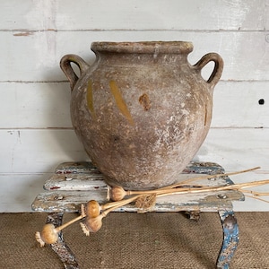 May include: A weathered, round, brown terracotta vessel with two side handles and a wide mouth. The exterior surface features a textured, mottled finish with faint yellow markings. The vessel is positioned on a small, distressed white and blue painted wooden stool, accompanied by several dried poppy seed pods lying across the stool surface.