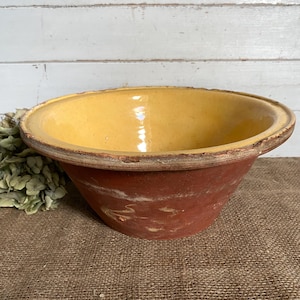 May include: A large, round, earthenware mixing bowl with a wide, flat rim. The interior is finished with a smooth, glossy, mustard-yellow glaze, while the exterior features a matte, unglazed, terracotta-red surface with visible wear and texture.