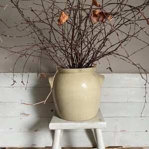 May include: A cream-coloured ceramic crock with two handles, filled with bare branches and a few dried leaves. The crock sits on a small, white, three-legged stool. The background is a white-painted wooden wall.