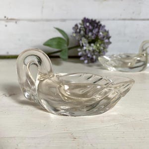 May include: A clear glass swan-shaped dish with a curved neck and a bowl-like body. The dish is on a white surface, with a second swan dish in the background. A purple flower and green leaves are also in the background.