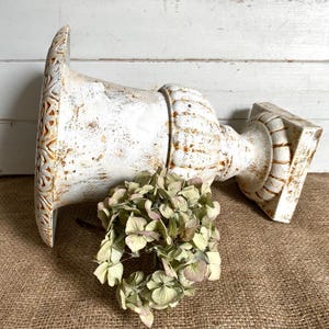 May include: A white urn with a distressed finish and brown accents, featuring a wide flared rim with a geometric pattern and a ribbed body on a square base, positioned horizontally next to a cluster of dried green and pale pink hydrangea flowers.