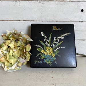 May include: A square black box featuring a painted floral arrangement of white bell-shaped flowers, yellow blossoms, and blue petals, with a small yellow bird depicted above the flowers. The box has a smooth, dark finish and a flat top surface.