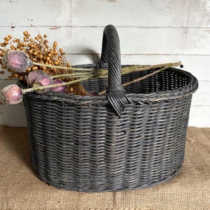 May include: A dark grey woven wicker basket with a curved handle. The basket contains dried flowers and seed pods. The basket is on a burlap surface. The background is a white wooden wall.