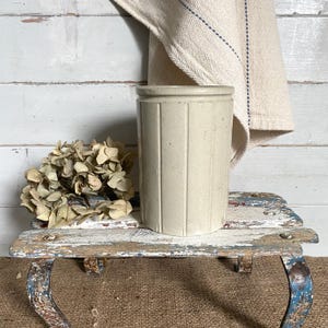 May include: A cylindrical off-white ceramic crock featuring vertical ribbed detailing along its exterior surface. The vessel has a smooth, matte finish and a simple, unadorned rim, presenting a neutral appearance suitable for various storage needs.