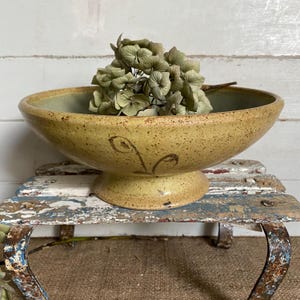 May include: A speckled mustard-yellow ceramic pedestal bowl with a wide, shallow basin and a flared base. The exterior features a hand-painted dark brown floral motif. A cluster of dried, muted green hydrangea blossoms rests inside the bowl.