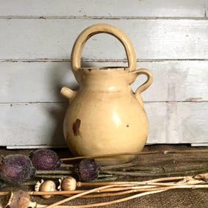 May include: A cream-coloured ceramic jug with a handle and a small spout. The jug has a rounded body and a loop handle. The surface has a rustic, aged appearance. The jug is approximately 20 cm tall.