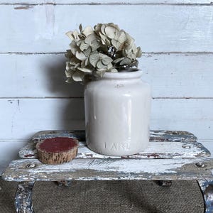 May include: A cream-coloured ceramic crock with a cylindrical body and a wide mouth, featuring the embossed text "1 LB" near the base. The crock contains a cluster of dried, pale beige hydrangea flowers and sits beside a small, round, red-painted cork.