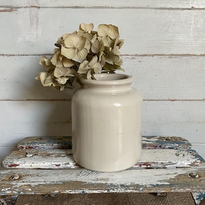 May include: A cylindrical cream-coloured ceramic crock with a smooth, glossy finish and a wide opening. A cluster of dried, light beige hydrangea flowers is placed inside the vessel, extending upwards from the top rim of the ceramic container.
