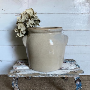 May include: A beige ceramic crock with a smooth, glossy finish and two small side handles. The vessel features a wide, rounded rim and a sturdy, cylindrical body. A cluster of dried, light brown hydrangea flowers is tucked inside the opening.