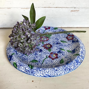 May include: A round ceramic plate featuring a dense blue and white floral pattern with accents of red and green. A single stem of purple lilac flowers with green leaves rests horizontally across the surface of the plate, partially covering the design.