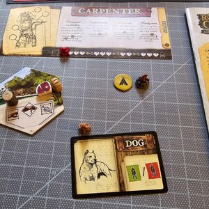 K&ouml;nnte beinhalten: Ein Spielbrett mit einer sechseckigen Kachel, die ein rotes St&uuml;ck Fleisch, einen braunen Baumstamm und eine Zeichnung eines Fisches zeigt. Eine Karte mit schwarzem Rand und dem Wort "DOG" in der Mitte zeigt einen wei&szlig;en Hund und zwei Figuren in Gr&uuml;n und Rot. Das Spielbrett befindet sich auf einer grauen Oberfl&auml;che.