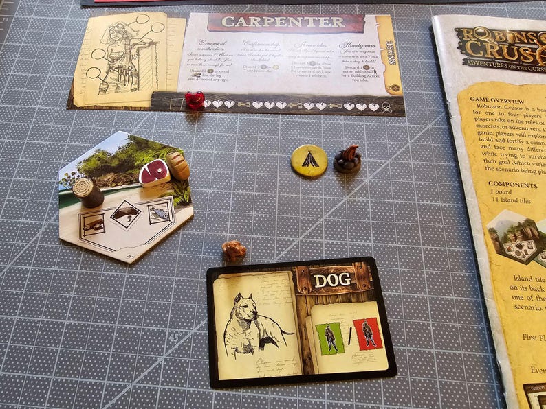 K&ouml;nnte beinhalten: Ein Spielbrett mit einer sechseckigen Kachel, die ein rotes St&uuml;ck Fleisch, einen braunen Baumstamm und eine Zeichnung eines Fisches zeigt. Eine Karte mit schwarzem Rand und dem Wort "DOG" in der Mitte zeigt einen wei&szlig;en Hund und zwei Figuren in Gr&uuml;n und Rot. Das Spielbrett befindet sich auf einer grauen Oberfl&auml;che.