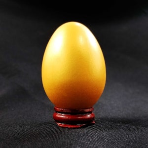 May include: A golden egg sits on a dark red wooden stand. The egg is smooth and shiny, with a slight pearlescent sheen.