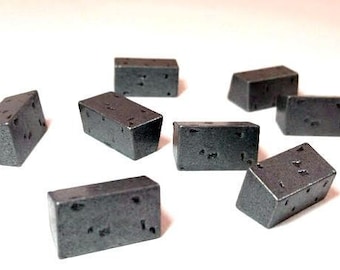 May include: A collection of eight small, dark gray, rectangular blocks with a slightly rough texture. Each block has several small, irregular holes on its surfaces. The blocks are scattered on a white surface.