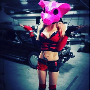 May include: A woman wearing a pink pig mask, red and black shorts, and a red and black top. She is posing with her hands on her hips.