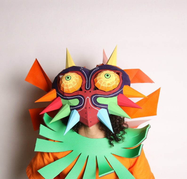 May include: A person wearing a papercraft mask of Majora's Mask from the Legend of Zelda video game series. The mask is red, green, blue, orange, and purple. The person is also wearing a green papercraft collar.