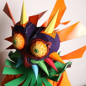 May include: A colorful paper mache mask of Majora's Mask from the Legend of Zelda video game series. The mask is made of paper and has a purple, orange, and green color scheme. The mask is worn by a person with dark hair.