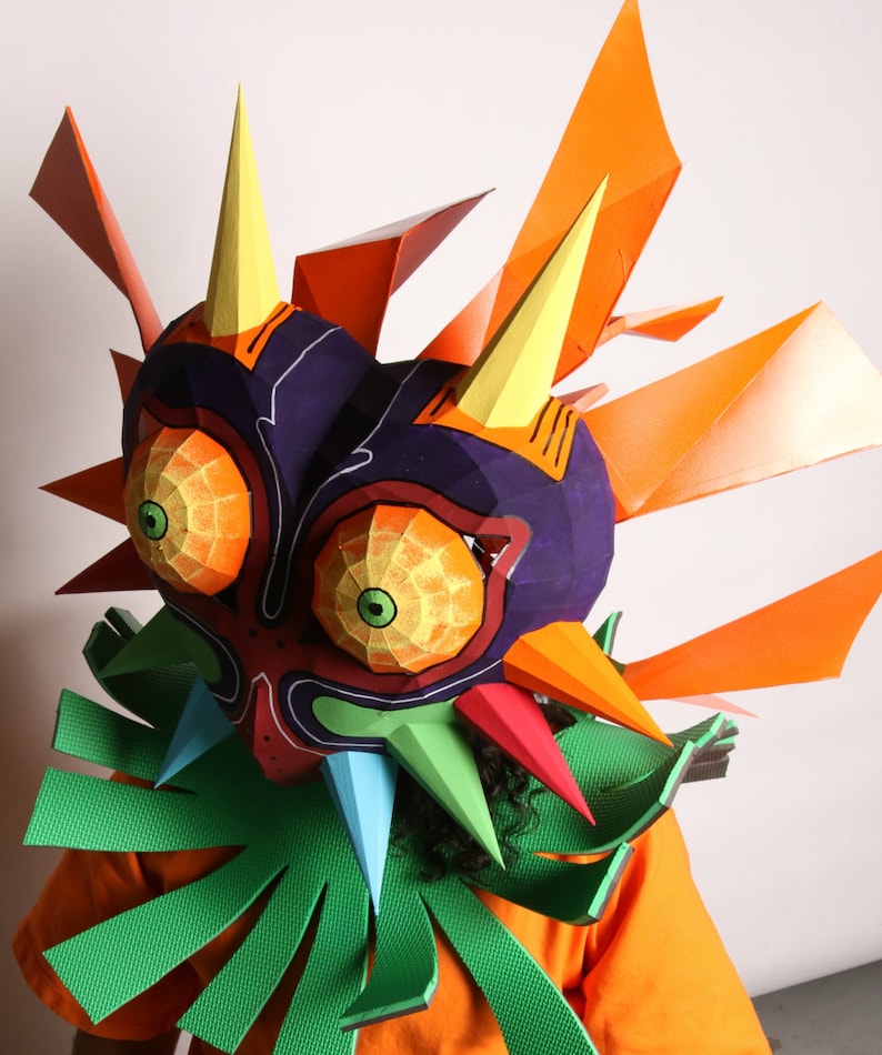 May include: A colorful paper mache mask of Majora's Mask from the Legend of Zelda video game series. The mask is made of paper and has a purple, orange, and green color scheme. The mask is worn by a person with dark hair.