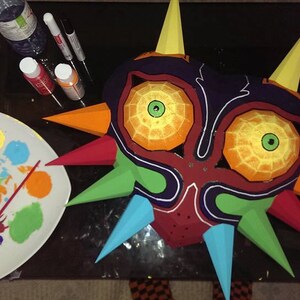 May include: A papercraft Majora's Mask from The Legend of Zelda: Majora's Mask video game is being painted. The mask is red, orange, yellow, green, blue, and purple. The mask is surrounded by paint pots and a paint palette.