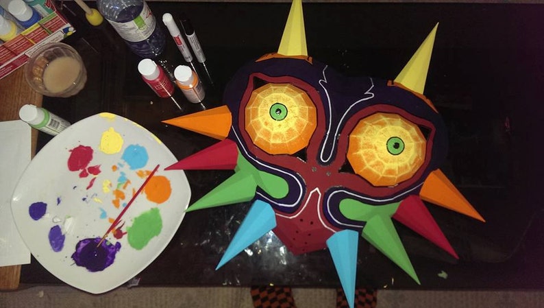 May include: A papercraft Majora's Mask from The Legend of Zelda: Majora's Mask video game is being painted. The mask is red, orange, yellow, green, blue, and purple. The mask is surrounded by paint pots and a paint palette.
